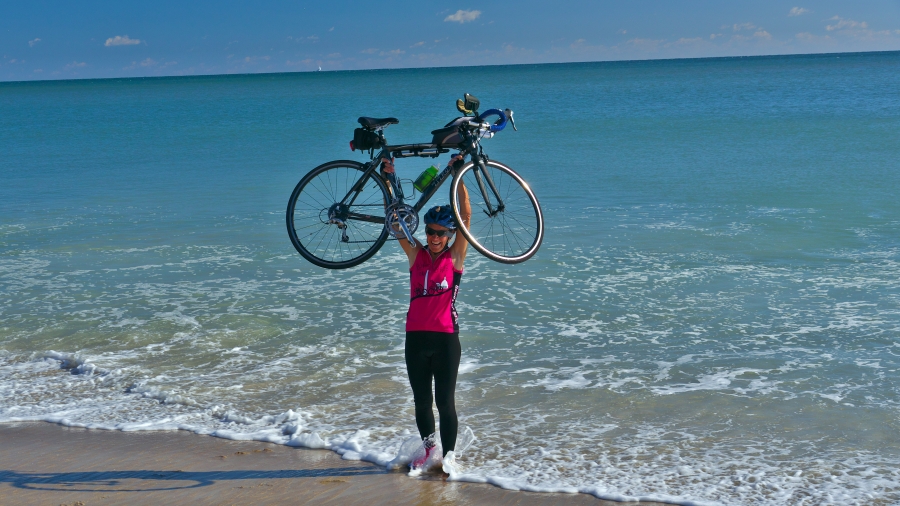 Lynn West Salvo ’71 hoists her bike over her head on the beach